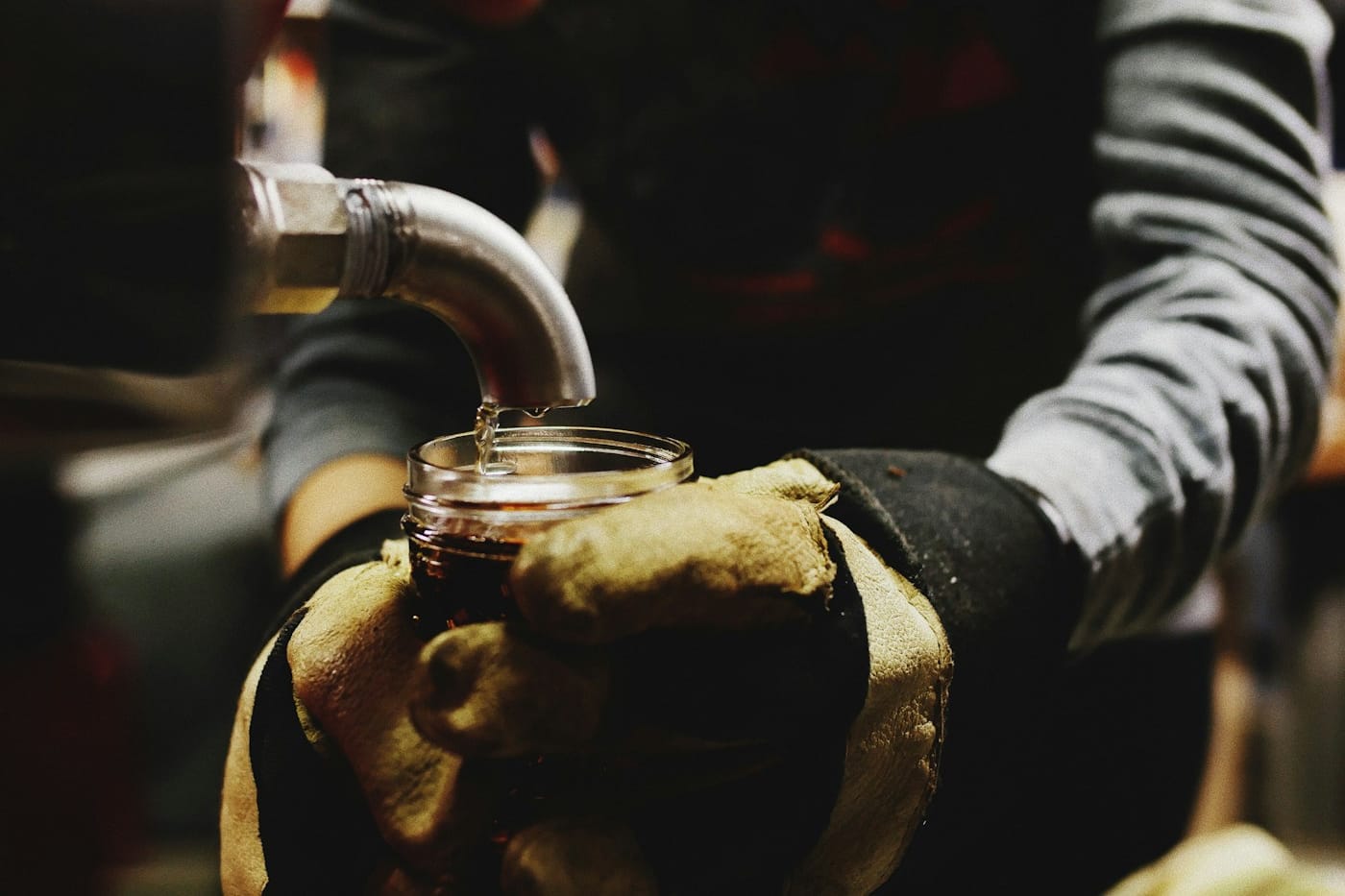 Gloved hands holding a jar beneath a metal tap, collecting maple sap.