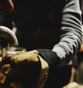 Gloved hands holding a jar beneath a metal tap, collecting maple sap.