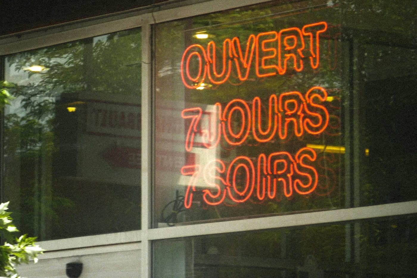 A red neon sign in a shop window reads “Ouvert 7 jours 7 soirs”, which means open seven days, seven evenings.