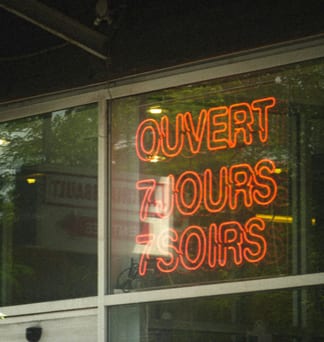 A red neon sign in a shop window reads “Ouvert 7 jours 7 soirs”, which means open seven days, seven evenings.