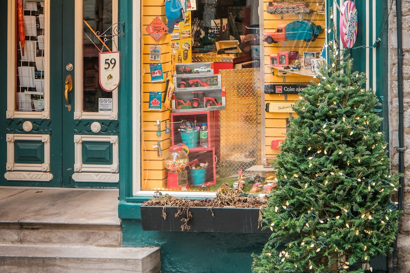 A small shop window full of vintage tin toys beside green doors and a lit evergreen tree on a narrow Québec City street.