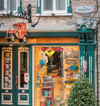 A small shop window full of vintage tin toys beside green doors and a lit evergreen tree on a narrow Québec City street.
